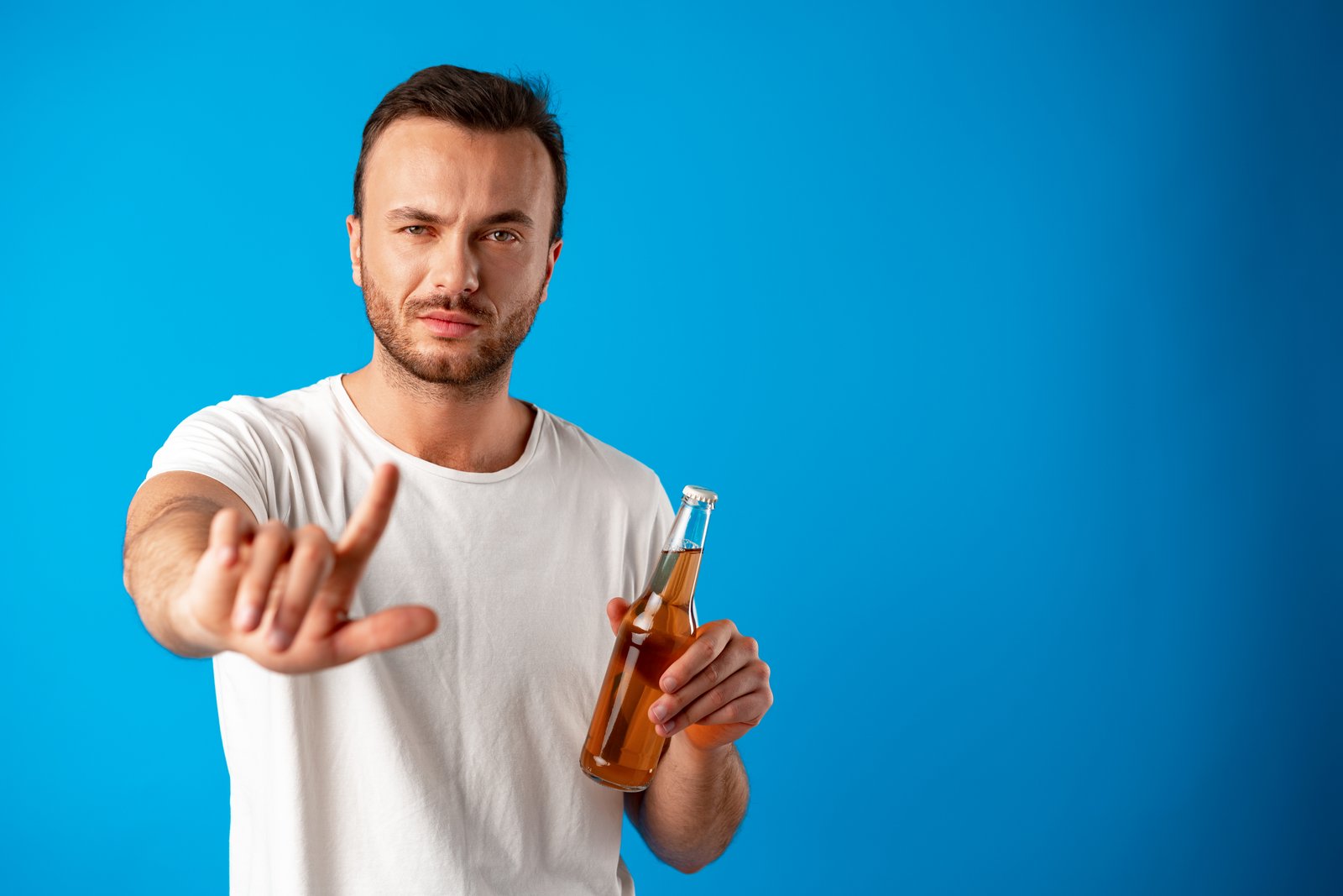 Young man holding a bottle of beer and shows a stop signal against blue background